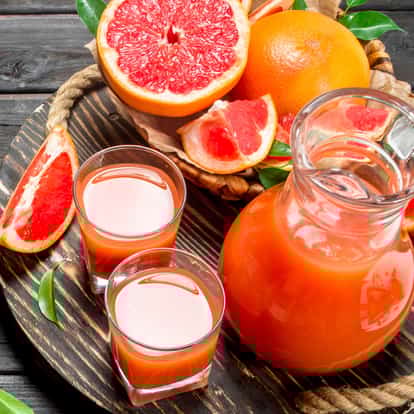 Ruby Grapefruit Juice in a glass pitcher with two short glasses and halved grapefruit on a round wooden board