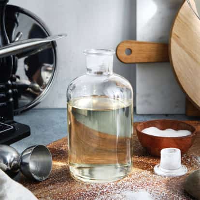 Sugar syrup in a glass bottle on a wooden board with a metal jigger and a small wooden bowl of sugar
