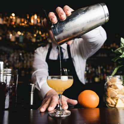 Suze in a coupe glass with liquid pouring from a metal shaker above, orange and jars on the bar