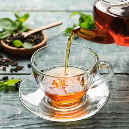 Tea pouring from a glass teapot into a glass teacup on a saucer with tea leaves and mint in the background