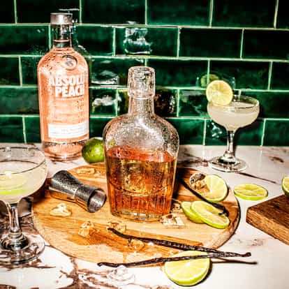 Vanilla Rum in a decanter on a board with vanilla pods and lime slices and an Absolut Peach bottle behind