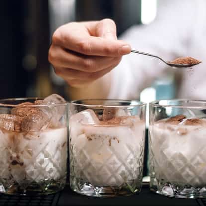 White Cacao Liqueur in three cut-glass rocks glasses with ice and cocoa powder dusted from a spoon