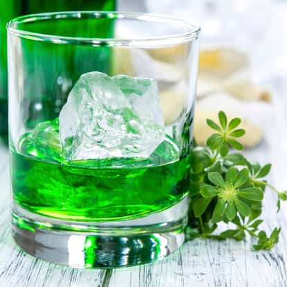 Woodruff Syrup in a rocks glass with a large ice cube and fresh woodruff sprig on white background.
