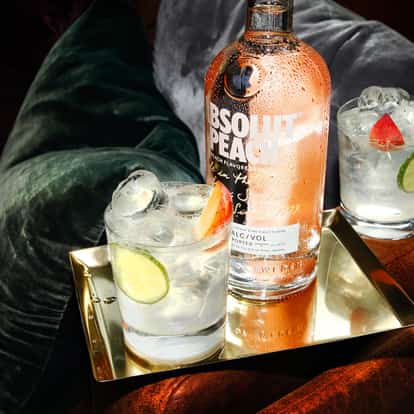 Absolut Apeach Soda bottle with condensation on a tray beside two iced rocks glasses with lime and peach slice