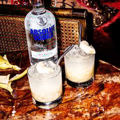 Absolut Banana Crush in two rocks glasses with crushed ice and banana slices beside a vodka bottle on a table