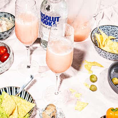 Bellini cocktail in a champagne flute next to a bottle of Absolut vodka and assorted bar snacks such as chips and olives.