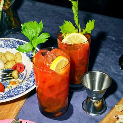 Absolut Bloody Mary in two highball glasses with lemon wheel and celery stalks beside a plate and grapes