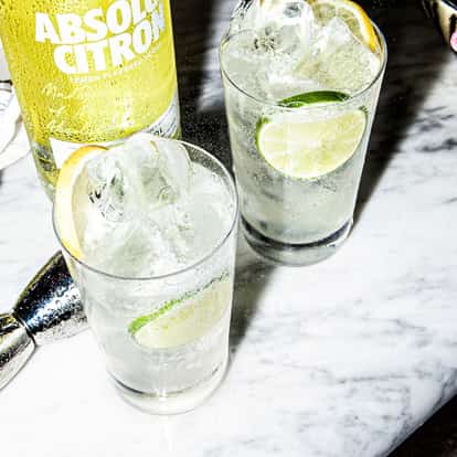 Lemon Vodka Soda two highball glasses with ice and lime slices beside an Absolut Citron bottle on a marble table