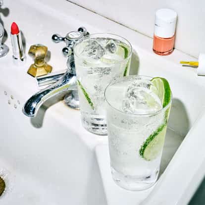 Absolut Fizzy Soda in two highball glasses with ice and lime wedges, on a sink with running faucet and nail polish