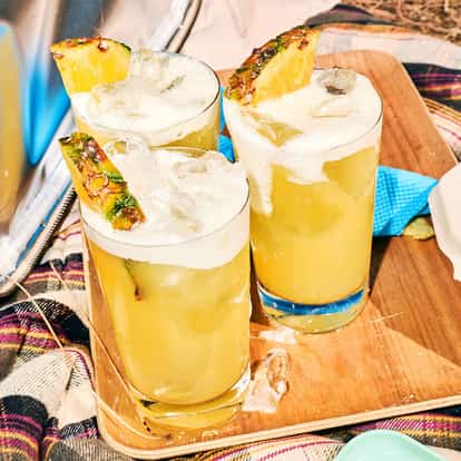 Absolut Fluffy Pineapple in three highball glasses topped with foam and pineapple wedges on a wooden board