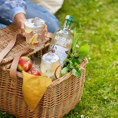 Absolut Juice Apple and Soda in two iced jars with apple slices and a bottle in a wicker picnic basket on grass