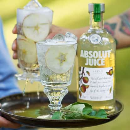 Absolut Juice Apple Spritz served in two stemmed glasses with ice and apple slices on a tray with a bottle