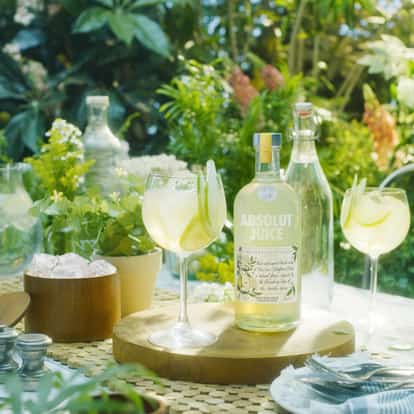 Absolut Juice Pear and Elderflower Shandy in two stemmed wine glasses with a bottle on a round wooden board