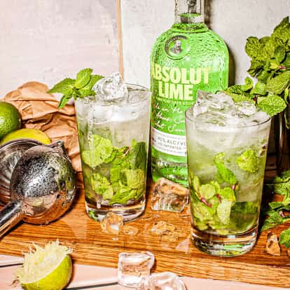 Absolut Lime Mojito in highball glasses with mint sprigs and ice, bottle and lime halves on wooden board