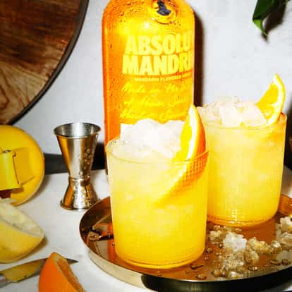 Absolut Mandrin Crush in two rocks glasses with crushed ice and orange wedges beside a bottle and mandarins on marble
