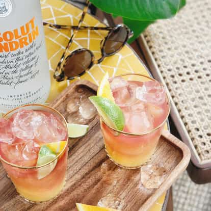 Absolut Mandrin Madras in two rocks glasses on a wooden tray with lime and orange wedges and a bottle visible