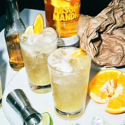 Absolut Mandrin Punch in 3 glasses with ice and orange and lime wedges and a bottle behind on white surface