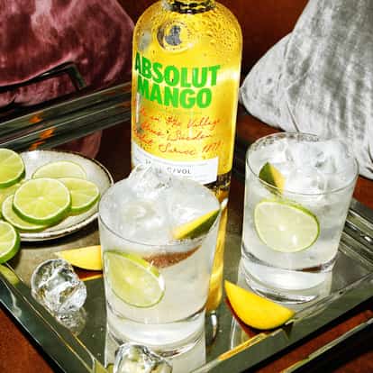 Two vodka sodas with lime in rocks glasses beside an Absolut Mango bottle on a mirrored tray.