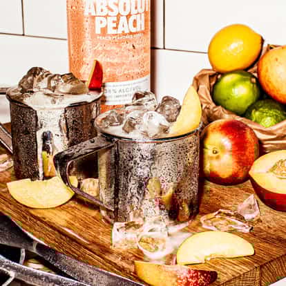 Absolut Peach Mule in three mule mugs with ice on a board with peach slices and a bottle in background
