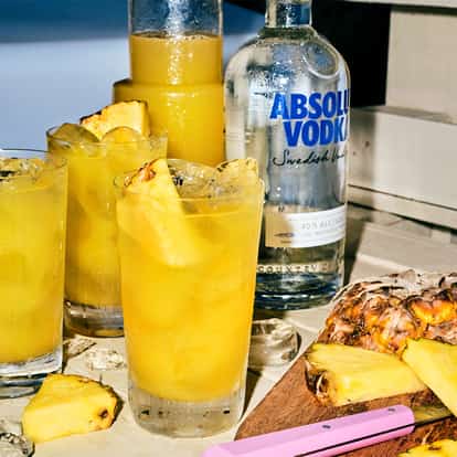 Absolut Piña in three highball glasses garnished with pineapple wedges and a vodka bottle on a wooden bench.