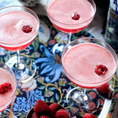 Four raspberry martinis in coupe glasses garnished with raspberries and an Absolut bottle behind.