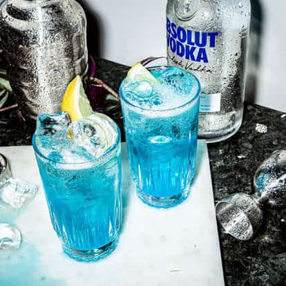 Absolut True Blue in two highball glasses with lemon wedges and ice on marble with a vodka bottle and jigger
