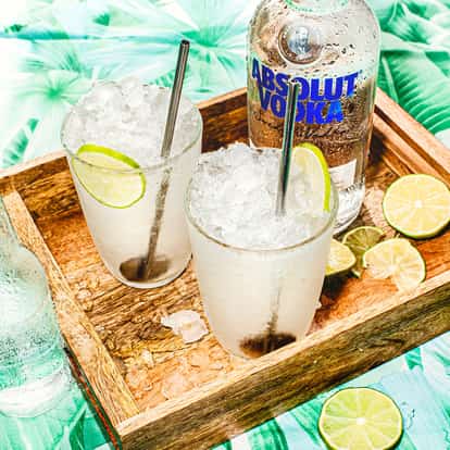 Absolut Twist served in two glasses with crushed ice and lime wheels on a wooden tray with a vodka bottle