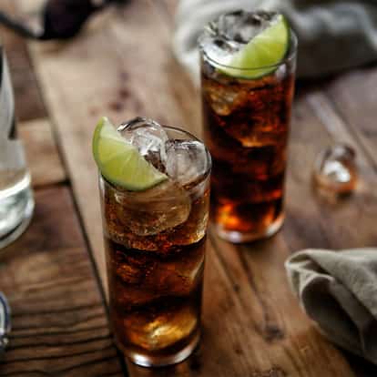 Two mixed drinks in highball glasses garnished with lime, beside a bottle of Absolut Vanilia on a wooden table.