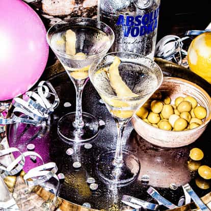 Festive party table with balloons, streamers, party hats and two martini glasses containing a Vodka Martini made with Absolut Vodka.