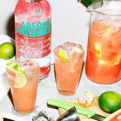 Absolut Waterlemon served in two highball glasses with ice and lime wedges and a pitcher on white background