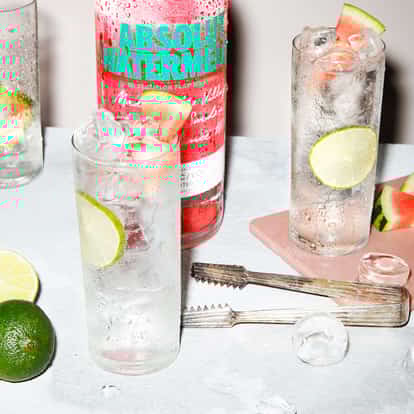 Absolut Watermelon and Soda cocktail next to a bottle of Absolut Watermelon and two limes.