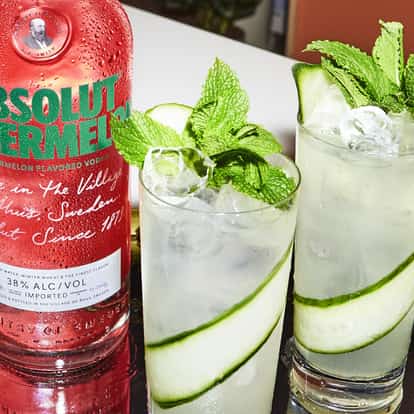 Two Absolut Watermelon Lemonade cocktails garnished with mint leaves and cucumber next to a bottle of Absolut Watermelon.