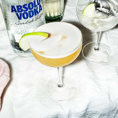 Apple Strudel Martini in a coupe glass garnished with an apple slice beside an Absolut Vodka bottle on white background.
