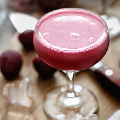 Beets Martini in a coupe glass with a frothy head on a wooden board beside beet halves and ice cubes
