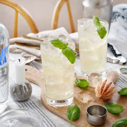 Two highball glasses of basil cocktail with basil leaves and ice on a board beside an Absolut vodka bottle.
