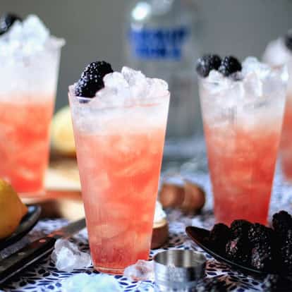 Four beautiful Blackberry Punch cocktails in tall glasses filled with lots of ice and garnished with fresh blackberries. In the background is a bottle of Absolut Vodka and several fresh lemons.