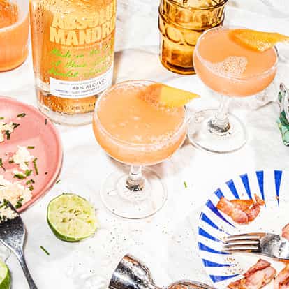 Blood Orange Cosmo served in two coupe glasses with orange twists beside an Absolut Mandrin bottle.