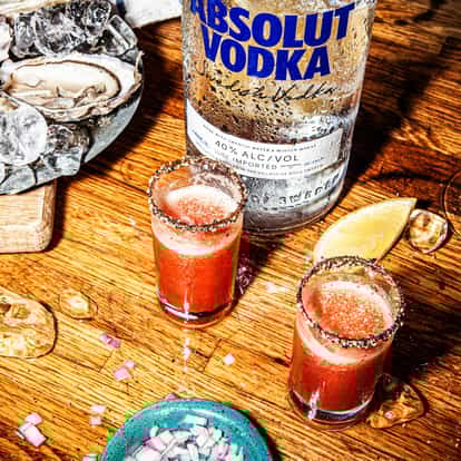 Two Bloody Mary Shots in two shot glasses with salted rims sat beside a bottle of Absolut Vodka, oysters and a lemon slice.