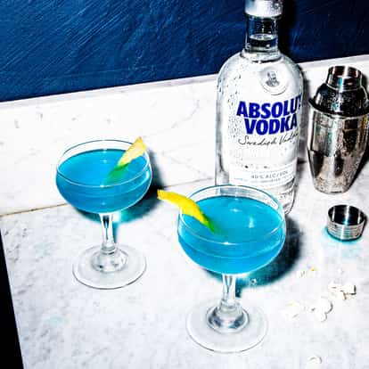 Two Blue Cosmopolitan cocktails garnished with lemon zest, next to a bottle of Absolut Vodka and a boston shaker.