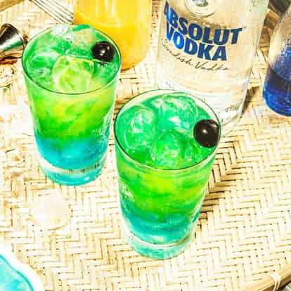 Blue Hawaiian in a highball glass with ice and a cherry garnish next to an Absolut Vodka bottle on a woven tray