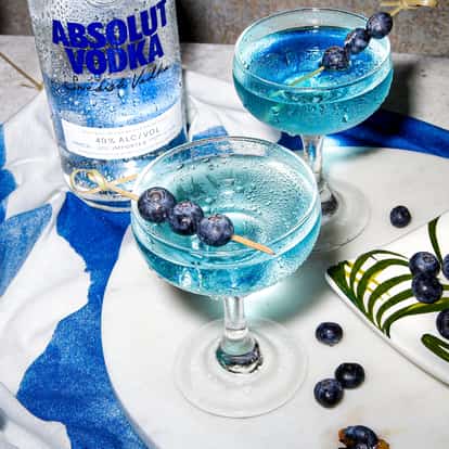 Blueberry Martini in coupe glasses with Absolut Vodka bottle and blueberry skewers on a marble board