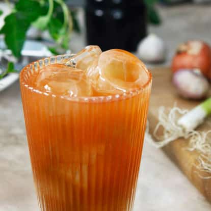 Bull Shot served in a ribbed highball glass with large ice spheres next to spring onions and shallots on a wooden board