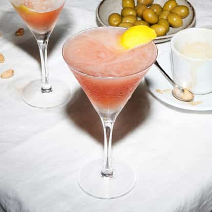 Pink cocktail in a martini glass with a lemon twist garnish on a white tablecloth.