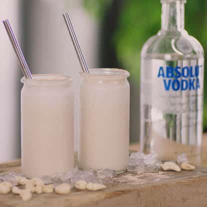 Cashew Hardshake in two glass jars with straws on wood with ice and cashews beside an Absolut Vodka bottle