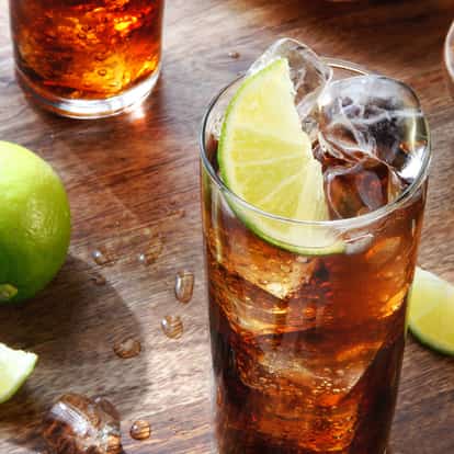 Cuba Libre in a highball glass with ice and a lime wedge on a wooden board