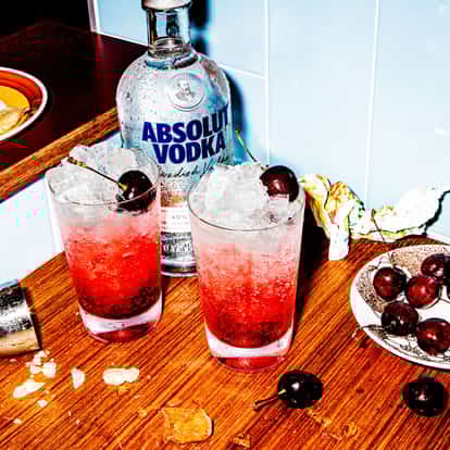 Dirty Shirley in two highball glasses with ice and cherries beside an Absolut Vodka bottle on a wood counter