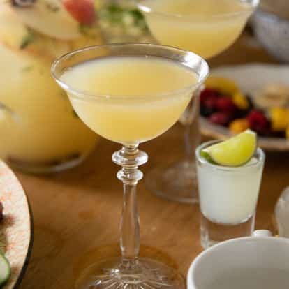A pale yellow cocktail in a stem coupe with a small lime-garnished shot on a wooden table.
