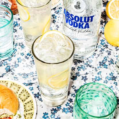 Elderflower Collins in a Collins glass with lemon slice and ice next to an Absolut vodka bottle on a floral cloth