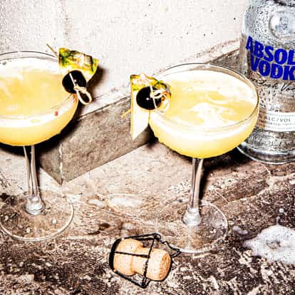 Flirtini served in coupe glasses garnished with pineapple and cherry, Absolut Vodka bottle on concrete floor