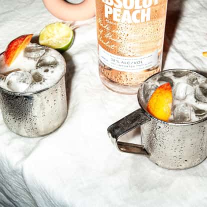 Moscow Mule varation, the Fuzzy Mule, made with Absolut Peach served in two aluminum mule mugs filled with ice and garnished with a peach wedge.